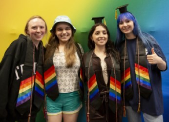 Four students pose with arms around each other, wearing rainbow-themed graduation regalia.