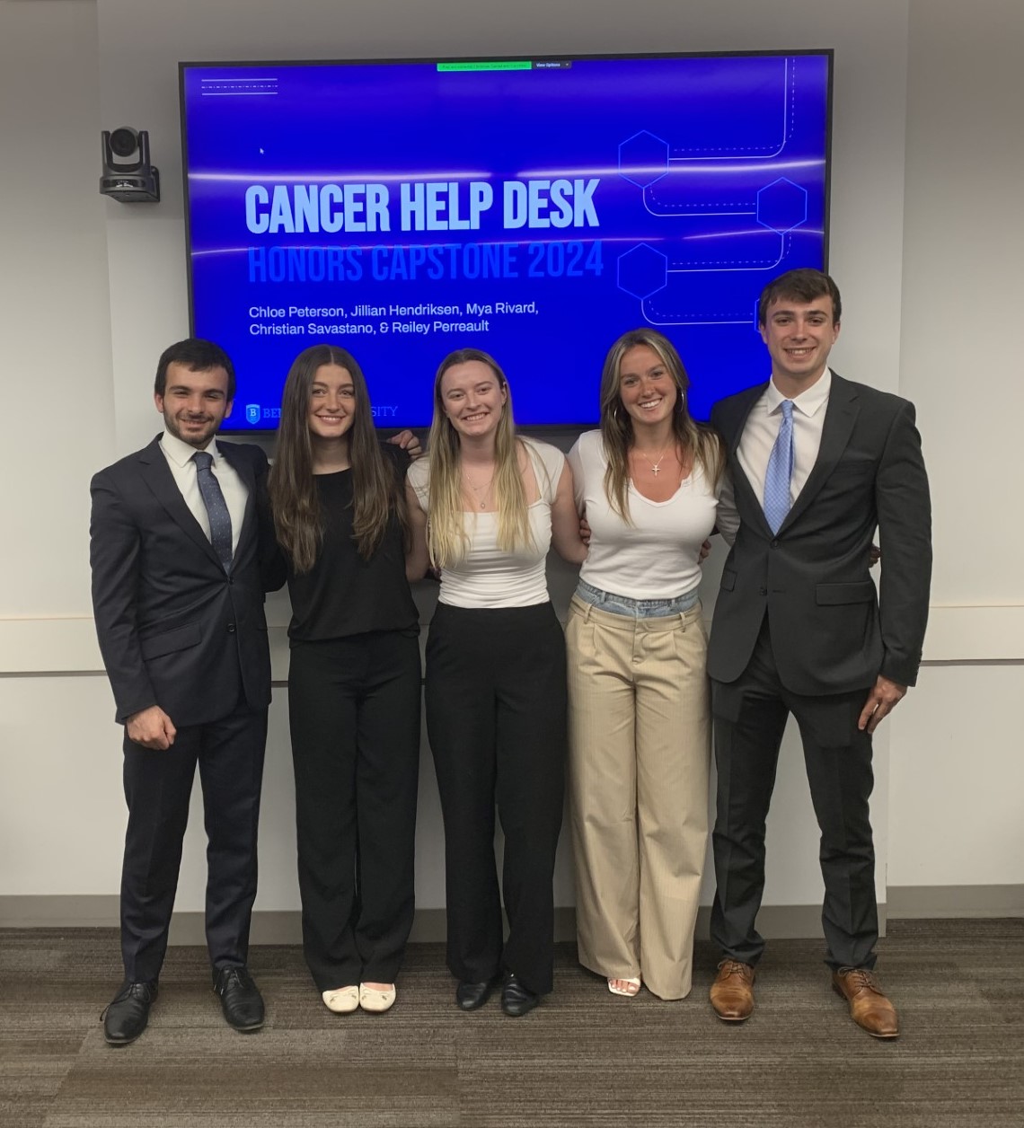 Bentley students (from L to R) Christian Savastano, Chloe Peterson, Mya Rivard, Jillian Hendriksen and Reiley Perreault pose in front of an enlarged screen showing the a slide from their presentation to Cancer Help Desk.