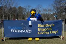 Flex, a costumed mascot in a Falcon suit stands near a banner for Bentley's Biggest Day of Giving 