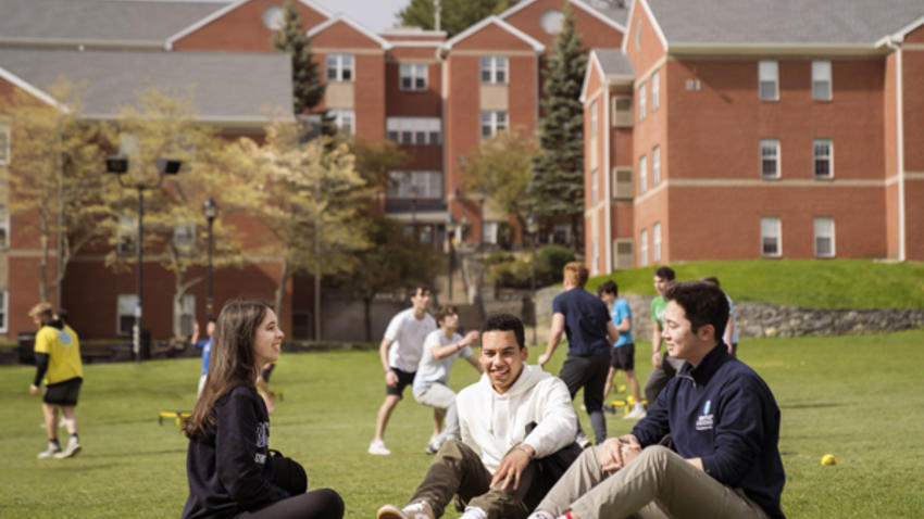 three students sit outside smiling on green grass with Bentley dorms in the background