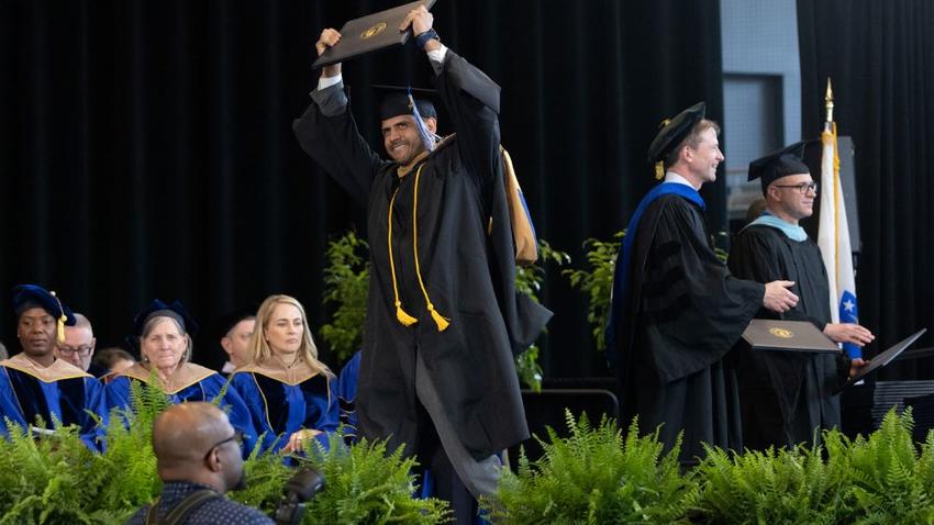 A graduate of the Bentley McCallum Graduate School of Business crosses the stage with diploma in the air during commencement
