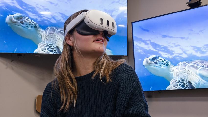 Caroline Chang ’24 sits in the foreground wearing VR googles; in the background, two large screens show identical images of what Caroline is seeing on the headset: a sea turtle swimming underwater.