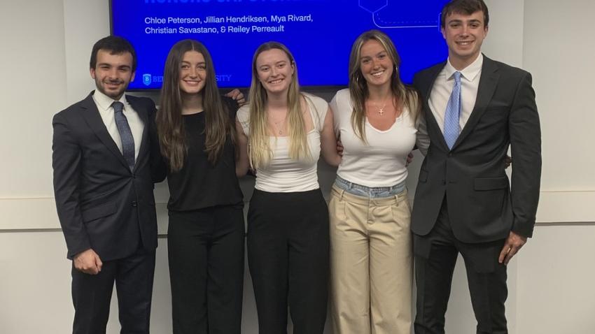 Bentley students (from L to R) Christian Savastano, Chloe Peterson, Mya Rivard, Jillian Hendriksen and Reiley Perreault pose in front of an enlarged screen showing the a slide from their presentation to Cancer Help Desk.