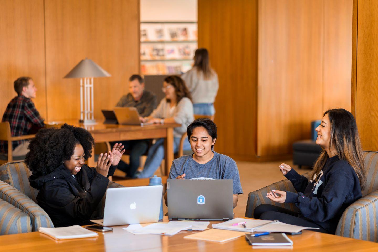 three students sit in the library talking and laughing